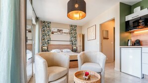 Ein modernes Hotelzimmer mit einem Bett, zwei Sesseln, einem kleinen Tisch mit Kerze, einer Küchenzeile inklusive Mikrowelle und einem großen Fenster mit Vorhängen.
