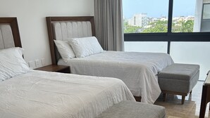 Une chambre d’hôtel avec deux lits, un pouf gris, une table de chevet en bois, un appareil de climatisation et une grande fenêtre donnant sur la ville.