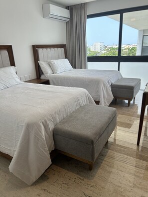 Ein Hotelzimmer mit zwei Betten, einem grauen Pouf, einem hölzernen Nachttisch, einer Klimaanlage und einem großen Fenster mit Blick auf die Stadt.