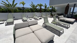 Un espace de détente aménagé sur le toit, avec plusieurs chaises longues et un canapé, entouré d’un mur blanc et de plantes tropicales.