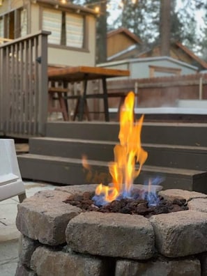 A fire pit with flames, surrounded by stone blocks, with a wooden deck and steps leading to a building in the background. - THE OHANA at BIG BEAR (Big Bear)