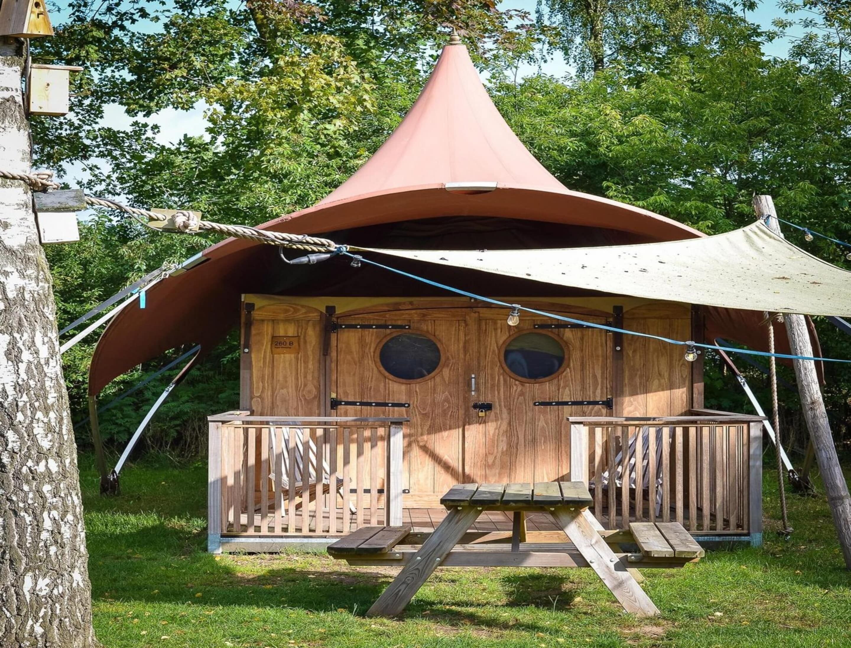 Eine Holzhütte mit einem Kegeldach, zwei runden Fenstern und einer Holzterrasse mit Picknicktisch.