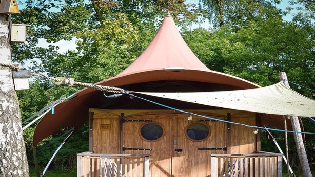 Eine Holzhütte mit einem Kegeldach, zwei runden Fenstern und einer Holzterrasse mit Picknicktisch.