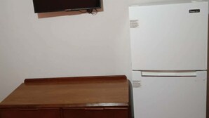 Una habitación con un banco de madera, un refrigerador blanco y un televisor montado en la pared.