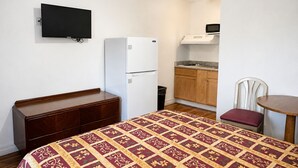 Una habitación pequeña y limpia con una cama, un aparador de madera, una kitchenette con microondas y un televisor fijado a la pared.