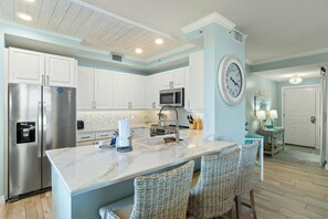 A modern kitchen with white cabinets, stainless steel appliances, a marble countertop, and a large wall clock. - Beach Colony East 2D (Pensacola)