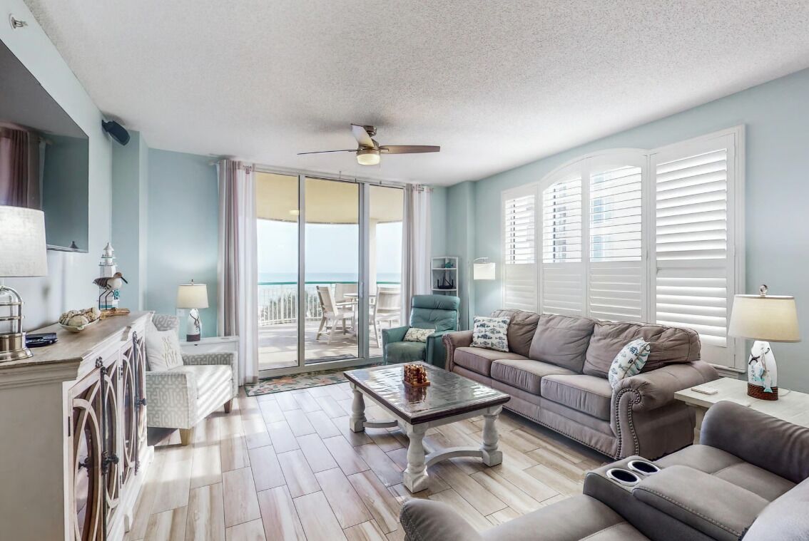 A living room with a view of the ocean, a ceiling fan, and a large window with blinds.