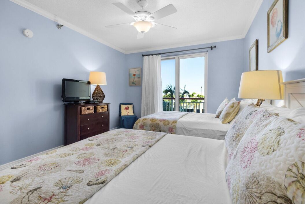A hotel room with two beds, a ceiling fan, a TV, a dresser, and a window with a view of palm trees.