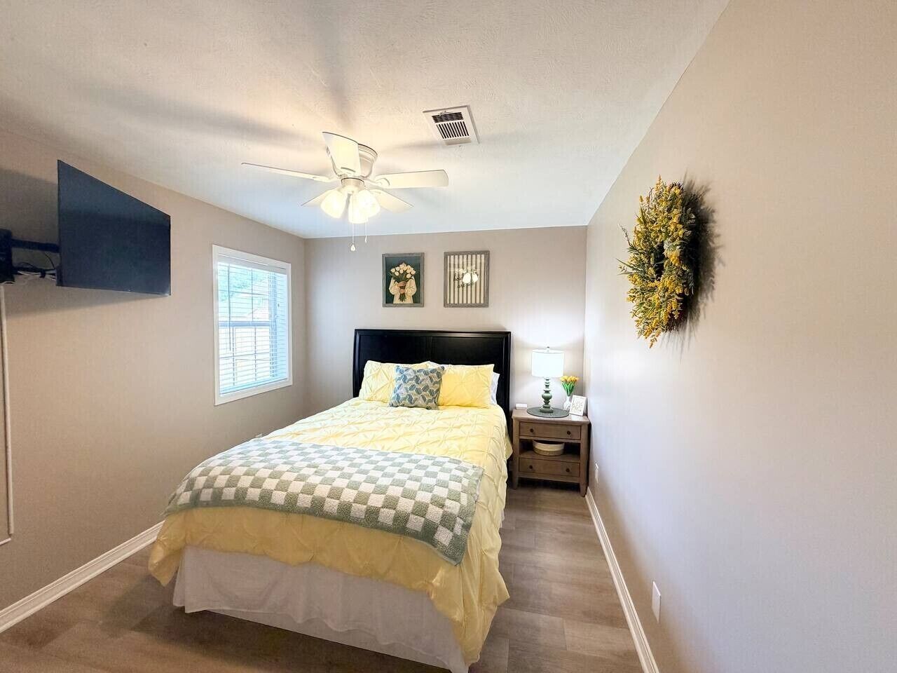 Une chambre à coucher comprenant un lit, une table de chevet, un téléviseur fixé au mur, un ventilateur de plafond, une fenêtre et une couronne décorative.