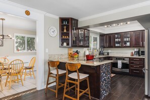 A modern kitchen with dark wood cabinets, a central island with bar stools, a built-in oven, and a dining area with wooden chairs.