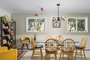 A dining area with a glass table surrounded by wooden chairs, a bookshelf with books and decorative items, and a ceiling light fixture.