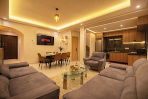 A modern living room with a glass coffee table, grey sofas, a dining area with a wooden table and chairs, a kitchen with wooden cabinets, and a mounted television displaying 'NETFLIX'. - Charming, family-friendly 2-bedroom apartment with AC in Marrakesh Targa (Marrakesh)