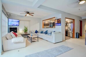 Una sala de estar moderna con sofá, mesa de centro, televisión, ventilador de techo y cocina visible a través de un arco.