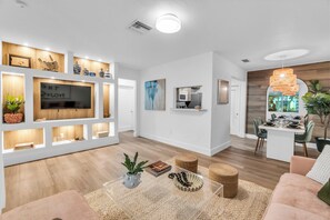 A modern living room with a TV, shelves with decorative items, a dining area with a table and chairs, and a kitchen area with a microwave. - Upscale 2-bedroom "Casita" 10  min away from Bal Harbor Beach, close to mall. (Biscayne Park)
