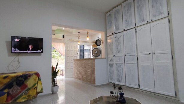 Quarto com televisão, planta em vaso, mesa com toalha colorida e guarda-roupa grande com várias portas.
