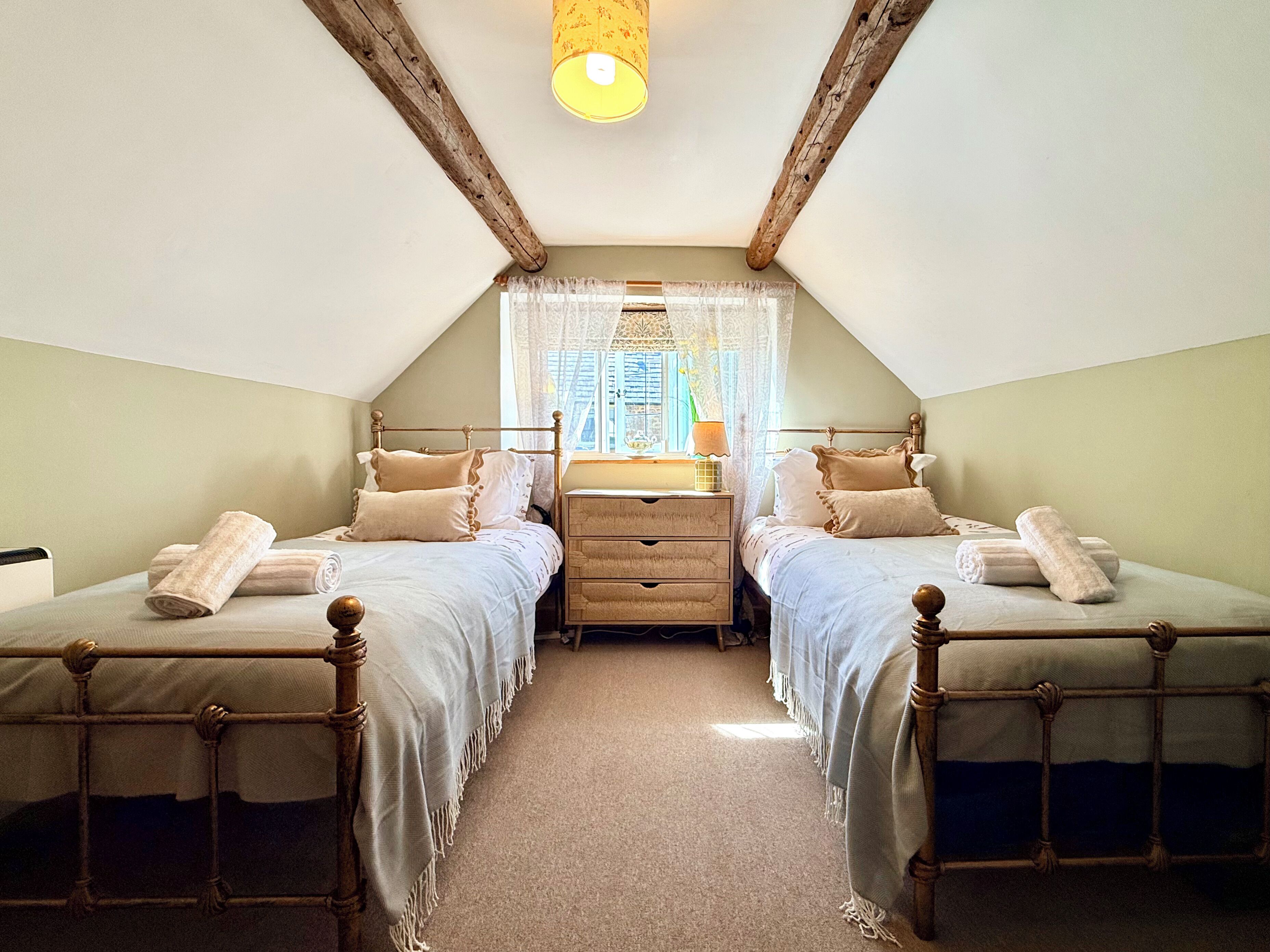 A bedroom with two single beds, a wooden dresser, a window with lace curtains, and a sloped ceiling.