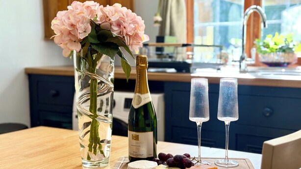 Et træbord med en champagneflaske, to glas, en vase med blomster og en træplanke med ost og druer.