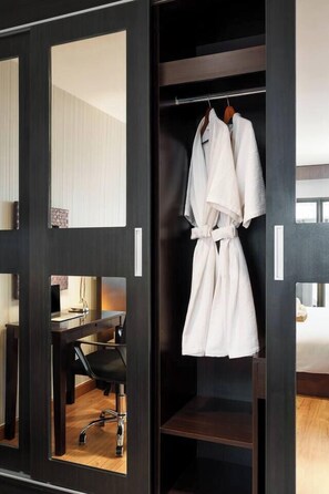Ein Hotelzimmer mit einem dunklen Holzschrank, einem Spiegel, einem aufgehängten Bademantel, einem Schreibtisch mit Stuhl und einem Bett.