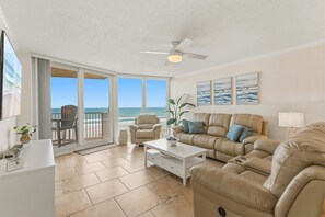 Ein Wohnzimmer mit einer Couch, einem Couchtisch und einem Fernseher. Es gibt einen Balkon mit einem Stuhl und einer Pflanze.