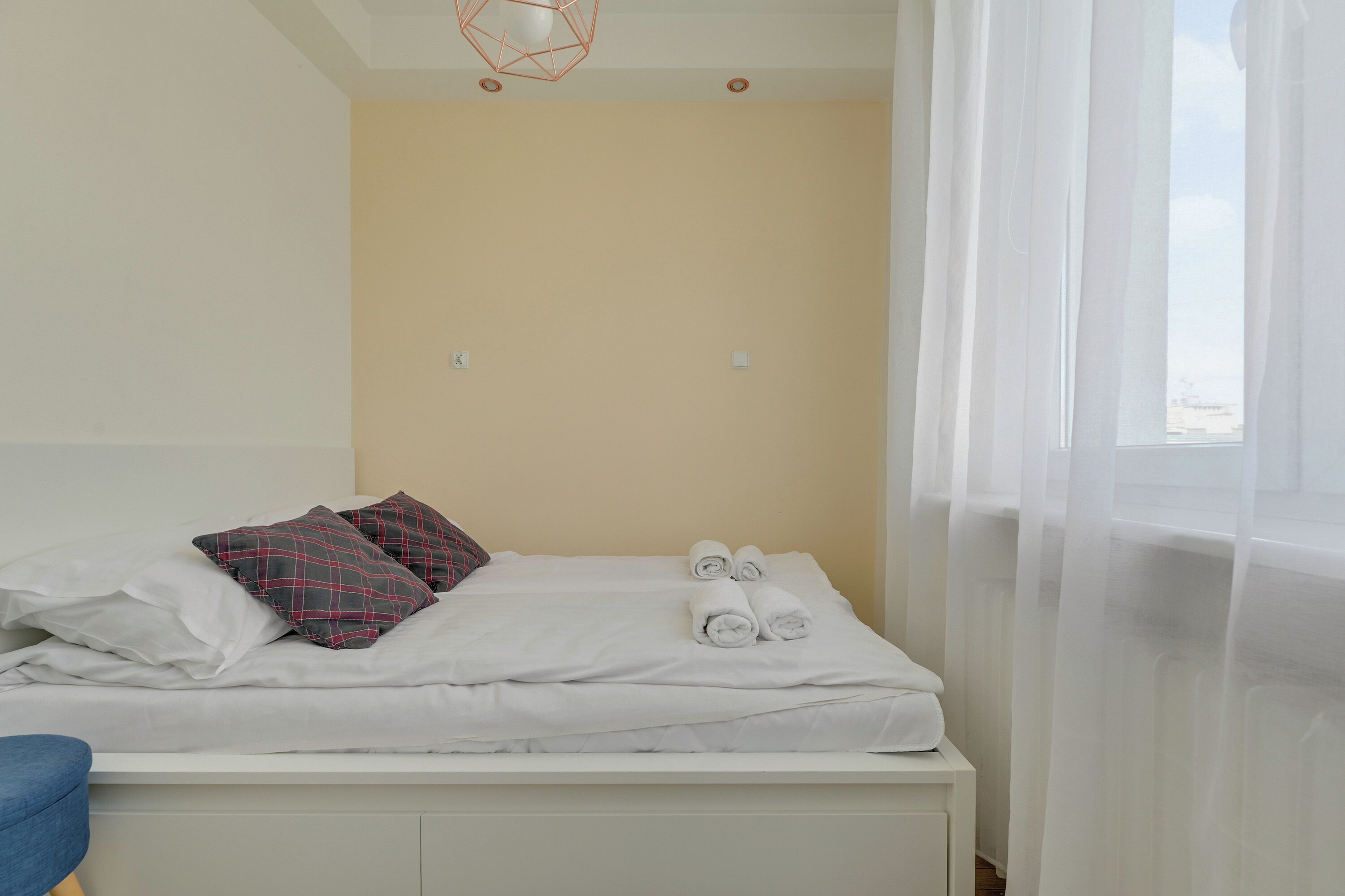 Een opgemaakt bed met kussens en opgerolde handdoeken, een blauwe stoel, en een raam met dunne gordijnen.