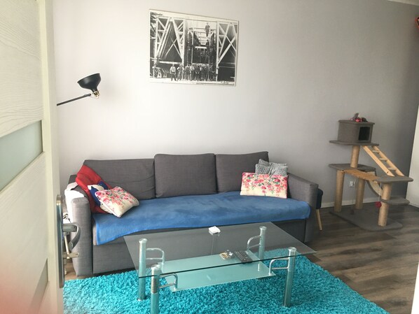 Ein Wohnzimmer mit grauem Sofa, einer blauen Decke, einem Glastisch, einer Katzenhöhle und einem Schwarz-Weiß-Foto an der Wand.