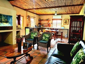 A living room with a sofa, two wicker chairs, a coffee table, a kitchen area with a dining table, and a bookshelf.