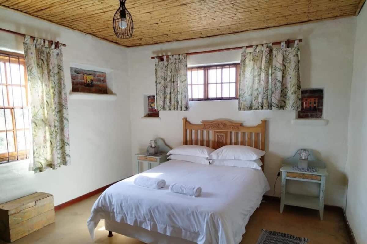 A bedroom with a wooden bed, white bedding, a nightstand, a small table, and a window with curtains.
