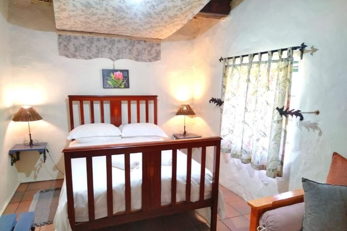 A bedroom with a wooden bed, two bedside lamps, a window with curtains, and a small table with a pillow.