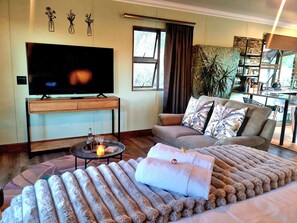 A living room with a flat-screen TV, a sofa with patterned cushions, a round coffee table with a candle, and a large window with curtains. - Fairfield Cottages (Ceres)