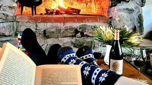 A fireplace with a pot, a lit fire, a book, a bottle of wine, and glasses on a table. - Fairfield Cottages (Ceres)
