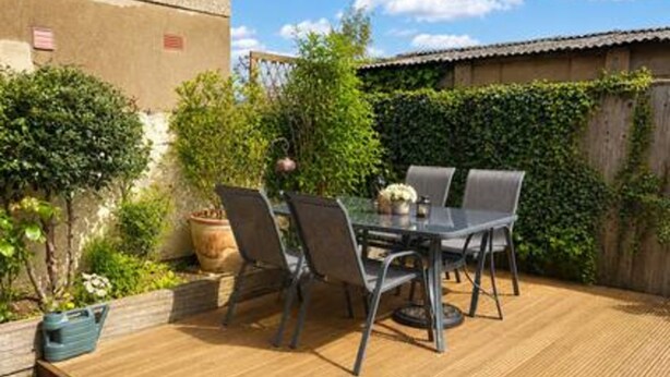 Un patio al aire libre con mesa y sillas, rodeado de follaje y una terraza de madera.