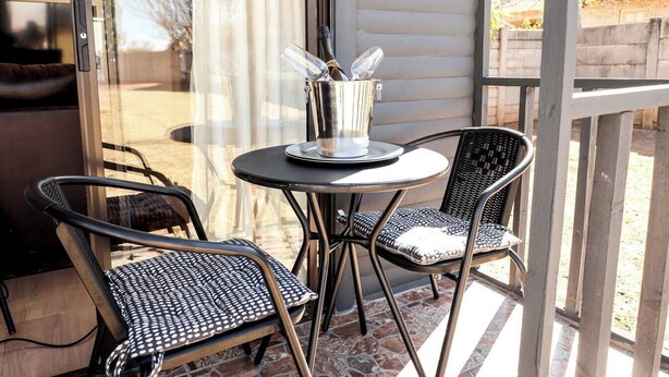 Un patio con una mesa y dos sillas, un recipiente de vidrio con una botella y vasos, y vista a una casa a través de una puerta de vidrio.