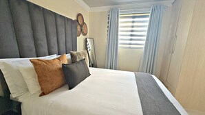 Habitación de hotel con una cama grande, cabecera, mesita de noche, lámpara y ventana con cortinas.