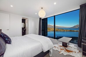 Une chambre avec un grand lit, une petite table, une vue sur un lac et des montagnes, et une porte donnant sur une autre pièce.
