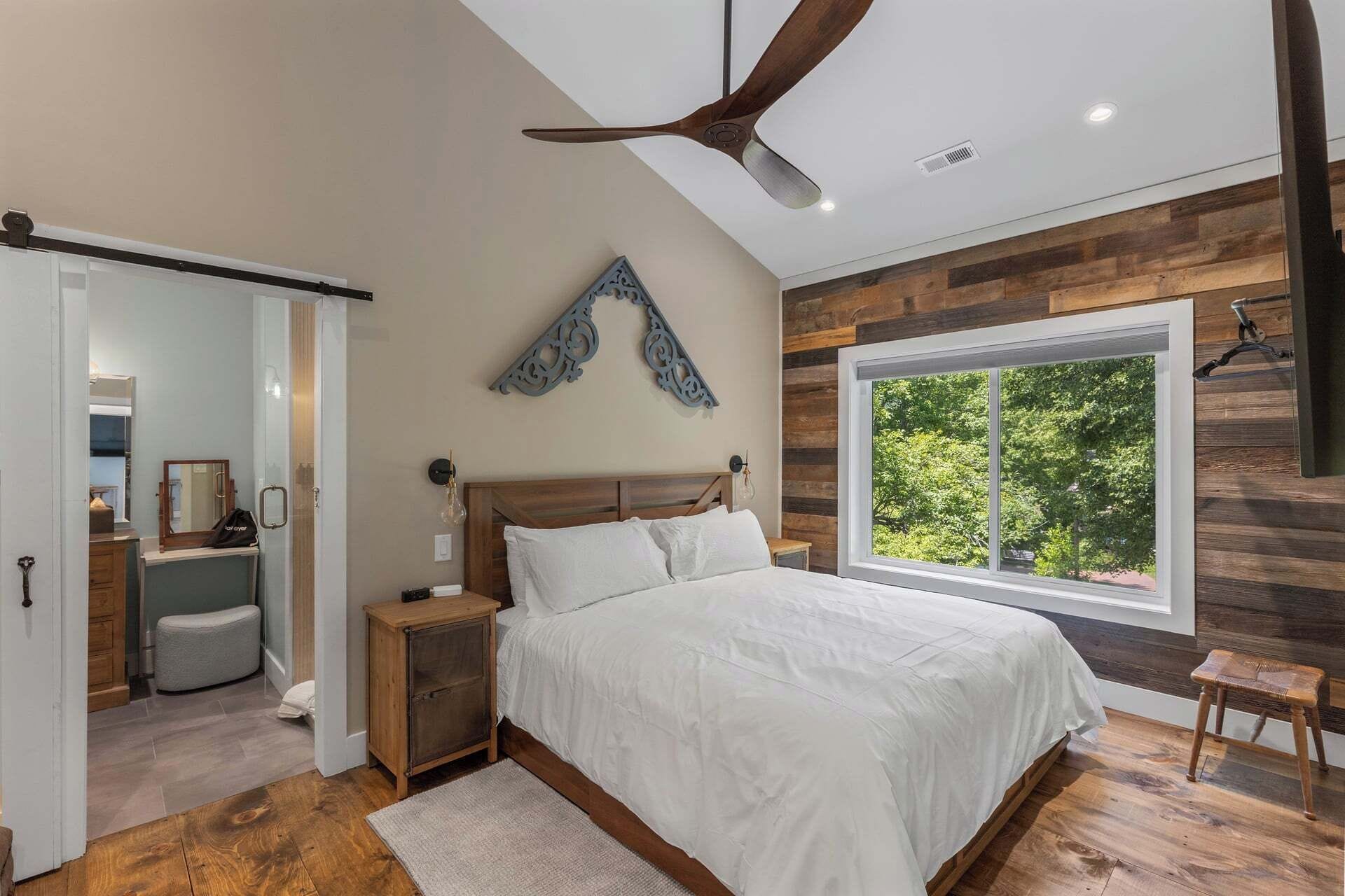A bedroom with a bed, a ceiling fan, a window with a view, and a wooden wall.