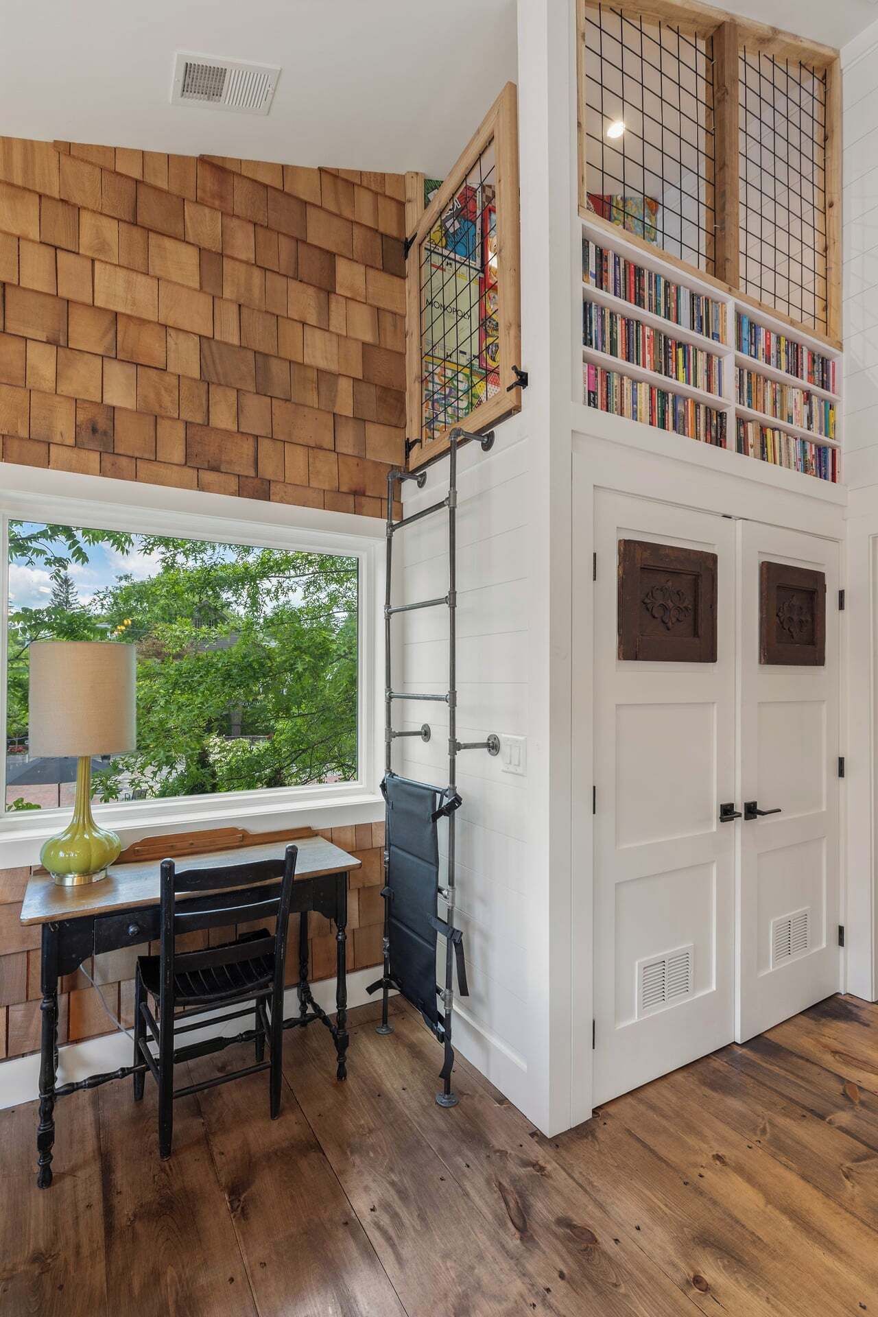 A room with a wooden wall, a window with a view, a table with chairs, and a bookshelf.