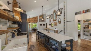 Cozinha com ilha central grande, prateleiras de madeira, luminárias pendentes e uma estante repleta de livros.