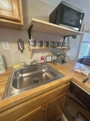 A kitchen with a stainless steel sink, wooden cabinets, a microwave, and a shelf with metal mugs.