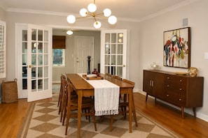 A dining area with a wooden table, chairs, a patterned rug, a chandelier, and a cabinet with decorative items. - Welcome to Brookside Mod Cottage: Spacious and group friendly! (Kansas City)