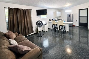 A living room with a couch, a fan, and a kitchen area with a refrigerator and a TV. - 9BR Home in Dorado PR | Near Beach & Groups (Dorado)