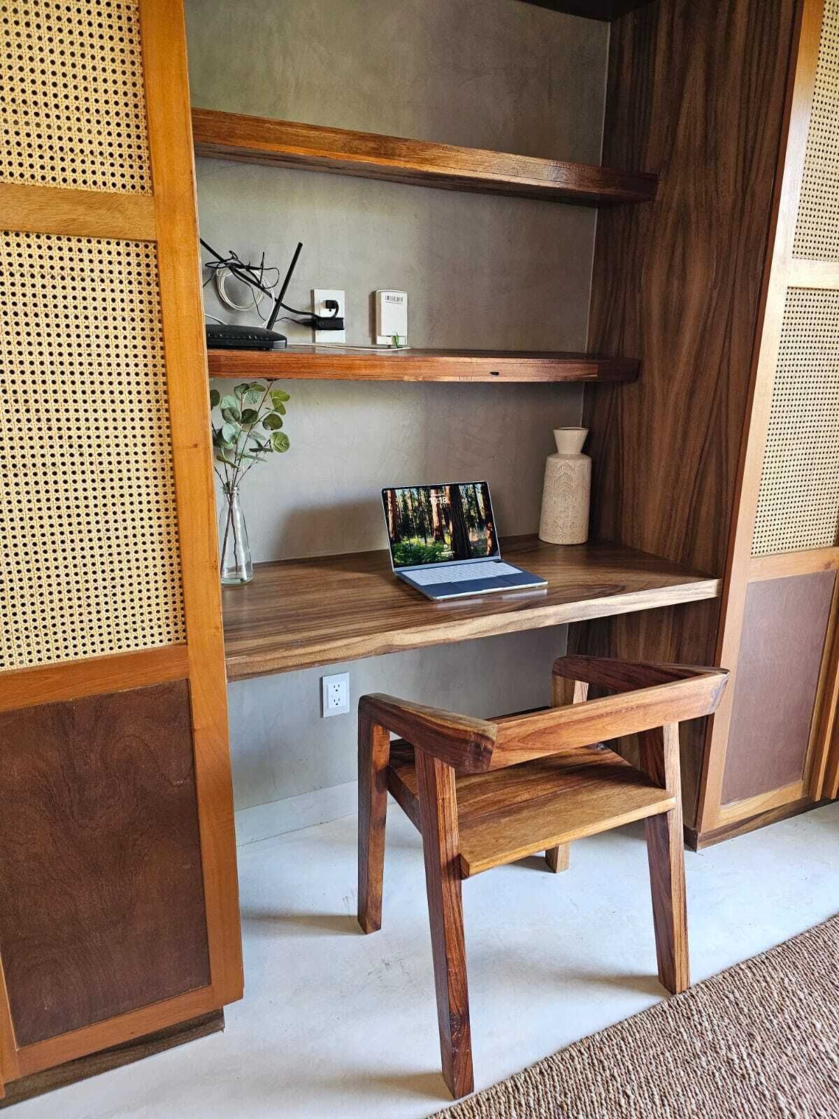 Un bureau en bois avec un ordinateur portable, un vase contenant des plantes vertes et une petite étagère sur laquelle se trouve un objet blanc.