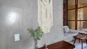 Ein Zimmer mit Wandbehagung aus Makramee, einem Holztisch, einer Pflanzvase und einem Korbstuhl.