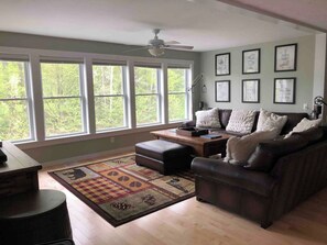 A spacious living room with a large window, a ceiling fan, a dark leather sofa, a wooden coffee table, and a patterned rug. - Trout River Lodge - Discount Jay Peak Lift Tix (Montgomery)