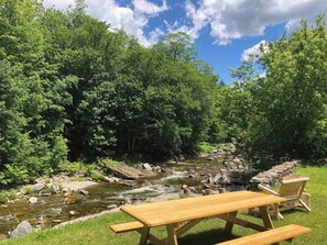 A picnic table and chairs by a river in a forested area.