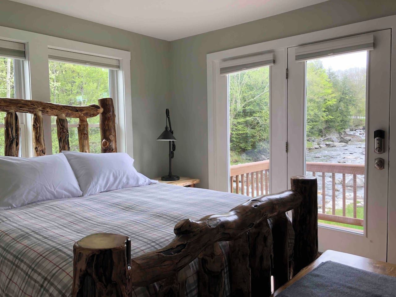 A bedroom with a large bed, a wooden headboard, a nightstand, a lamp, and a view of a natural outdoor area through a sliding door.