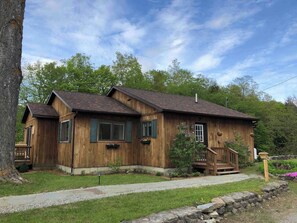 A wooden cabin with a porch, a stone wall, and a grassy yard.