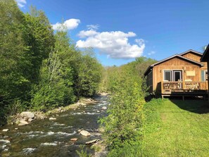 Una cabaña de madera con terraza, un río al frente y abundante vegetación alrededor.