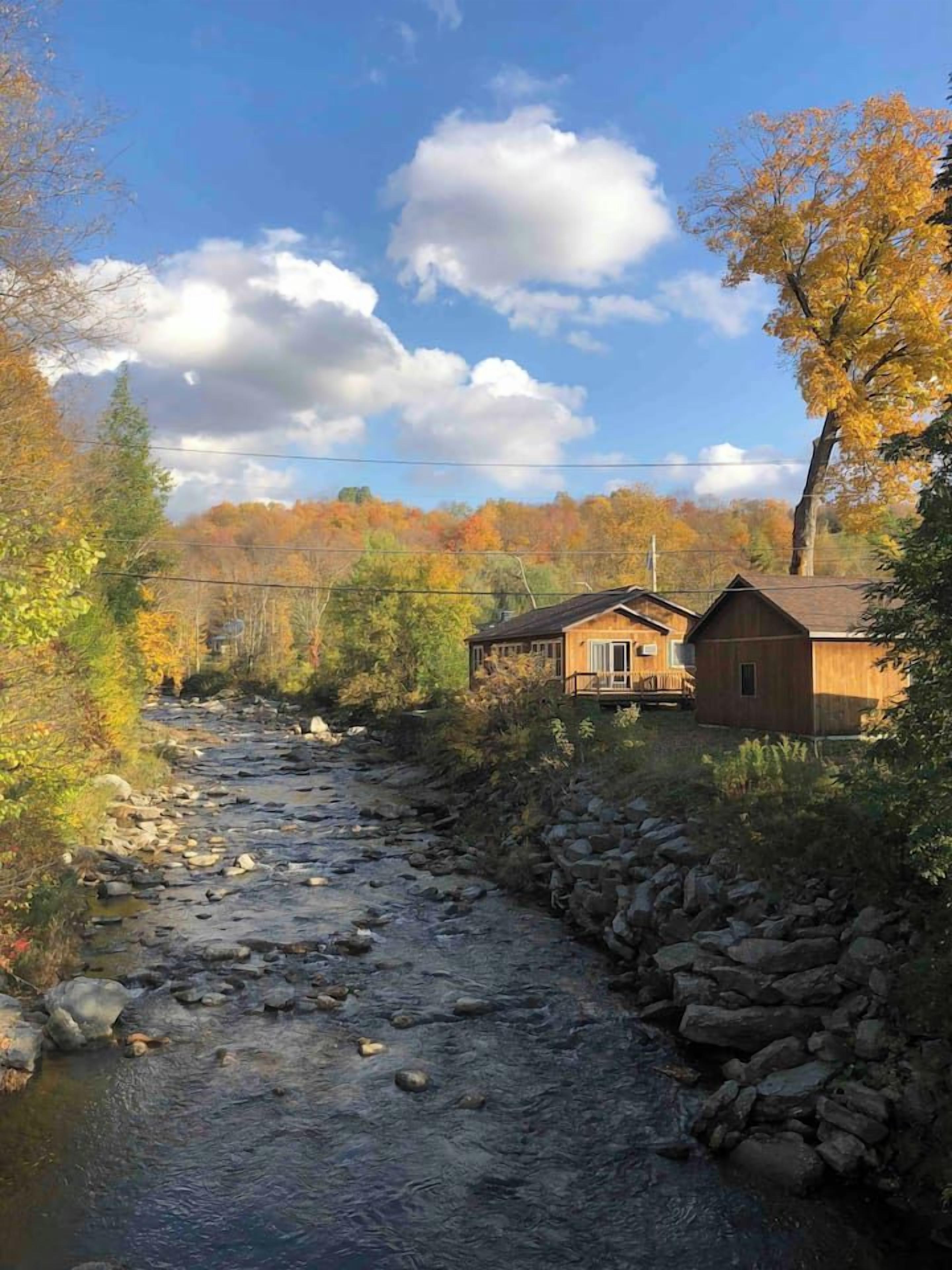 A rustic cabin with a stone foundation, a wooden deck, and a river flowing through a wooded area.