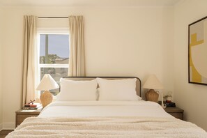 Una cama bien hecha con ropa de cama blanca, dos lámparas de noche, una mesita de mimbre con libros y una ventana con cortinas.