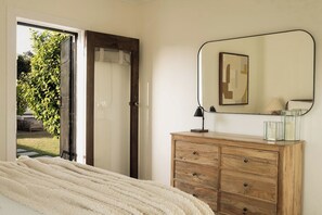 A bedroom with a bed, a wooden dresser, a mirror, and a door leading to a garden.